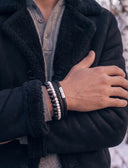 A close-up of a person wearing a stack of leather and beaded bracelets on their wrist, styled with a black sherpa-lined coat over a grey knit sweater.