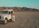 A woman leans against a vintage land rover in a desert setting