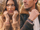 A split lifestyle image showing a woman in a brown knit turtleneck and a man in a brown shearling-lined jacket, both wearing stacks of gemstone beaded bracelets.