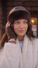 A woman wearing a brown fur hat, brown knit glove, a white fur coat, and colourful gemstone bracelets, standing in front of a rustic wooden cabin.