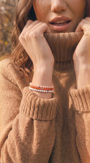 A close-up lifestyle shot of a woman wearing a brown knit turtleneck sweater and beaded bracelets on her wrist. The bracelet stack features Snow, Burnt Orange  and Ivory bracelets.